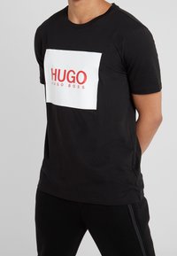 Sort bomulds t-shirt med en kontrastfarvet hvid rektangulær grafisk print med rød tekst "HUGO" og "HUGO BOSS" nedenfor. Korte ærmer, rund halsudskæring.