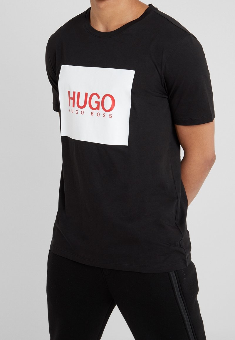 Sort bomulds t-shirt med en kontrastfarvet hvid rektangulær grafisk print med rød tekst "HUGO" og "HUGO BOSS" nedenfor. Korte ærmer, rund halsudskæring.
