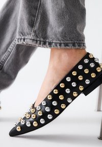 Zapato plano negro con tachuelas metálicas doradas y plateadas, diseño de punta afilada y textura lisa, combinado con unos jeans de mezclilla gris recortados.