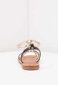 Sandali neri e metallici dorati con finitura testurizzata, cinturino alla caviglia regolabile e suola piatta in legno. Design con retro aperto.
