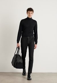 Melnā turtlenecka, slaida griezuma melni bikses un melni apavi. Nēsājot melnu somu, soļojot uz gaiši pelēka grīdas fona bez apdare.