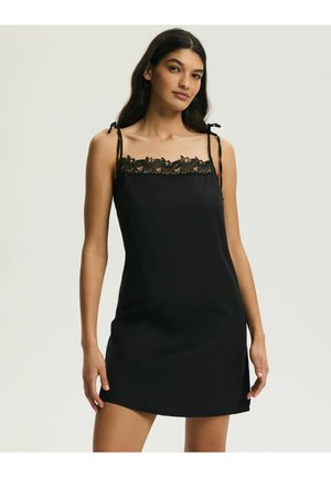 Kobieta ubrana w czarną, bezrękawową sukienkę na ramiączkach z koronkową lamówką przy dekolcie i wiązane ramiączka, stojąca na tle jednolitego tła.