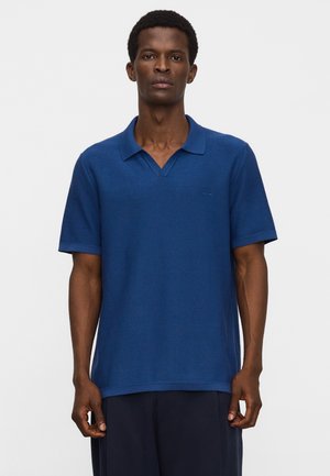 Mann trägt ein blaues Polohemd mit kurzen Ärmeln und dunkle Hosen, steht vor einem schlichten weißen Hintergrund.