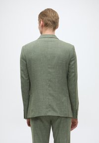 Grüne Leinenanzugjacke mit strukturiertem Finish, mit Revers und zwei Rückenschlitzen. Der Rücken des Models ist sichtbar und zeigt das taillierte Design.