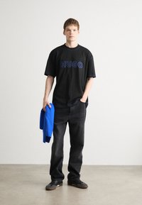 Melna pārmērīga T-kreklu ar zilu "HUGO" logo, kopā ar tumšām džinsām. Modelis tur zilu džemperi. Materiāls izskatās kā kokvilnas maisījums. Ikdienišķs piegriezums.