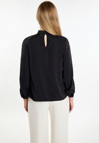 Schwarze Bluse mit langen Ärmeln und einem Schlüsselaugen-Detail am Rücken, mit Ballonärmeln und glatter Textur. Kombiniert mit hellen Hosen.