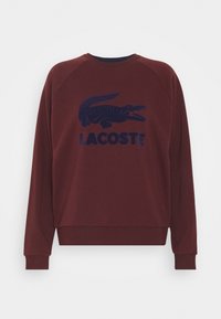 Sweatshirt bordeaux à manches raglan, avec un logo crocodile texturé bleu marine et le texte "LACOSTE" sur le devant. Tissu doux, coupe décontractée.
