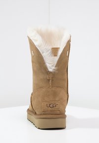 Botte en daim marron avec une doublure en peau de mouton, dotée d'une ouverture à l'arrière et du logo UGG sur le talon. Bout arrondi et semelle épaisse en caoutchouc.