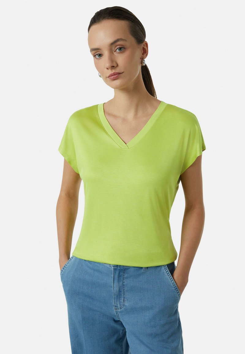 T-shirt w kolorze limonkowym z dekoltem w serek, krótkimi rękawami, miękką fakturą i luźnym krojem. Połączony z jasnoniebieskimi spodniami dżinsowymi, ręce w kieszeniach.