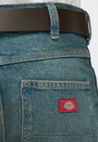 Τζιν παντελόνια εργασίας σε ανοιχτό μπλε χρώμα με ορθογώνια  τσέπη που φέρει κόκκινο διακριτικό Dickies. Καφέ δερμάτινη ζώνη με λεία υφή.