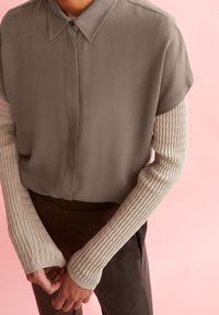 Brun kortärmad skjorta med krage, tillsammans med långa krämfärgade ribbade stickade ärmar. Bruna byxor fulländar looken.