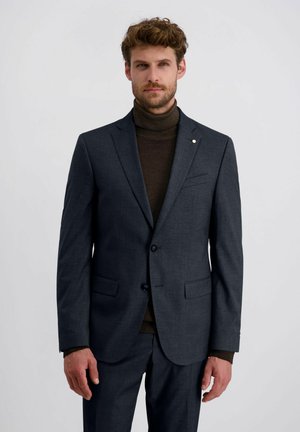 Mann mit lockigem Haar, der einen dunkelgrauen Anzug und einen braunen Rollkragenpullover trägt, steht vor einem schlichten weißen Hintergrund.