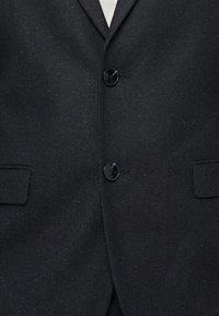 Μαύρο σακάκι με υφή, το οποίο διαθέτει δύο κουμπιά και δύο μπροστινές τσέπες. Διακριτικές μπλε κηλίδες προσθέτουν λεπτομέρεια χωρίς να είναι υπερβολικές.