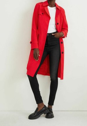 Un mannequin porte un trench-coat rouge vif sur un haut blanc, un jean skinny noir et des mocassins noirs épais, devant un mur uni.