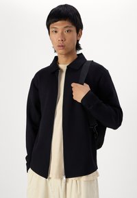 Veste noire zippée avec col, texture douce et manches longues. Portée par-dessus une chemise beige clair, assortie à un sac à dos noir.