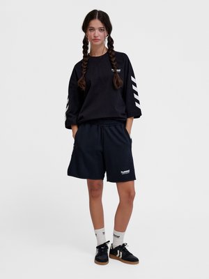 Jonge vrouw met lange vlechten die een zwart oversized sweatshirt en korte broek draagt, witte sokken en zwarte sneakers, staand met handen in de zakken.