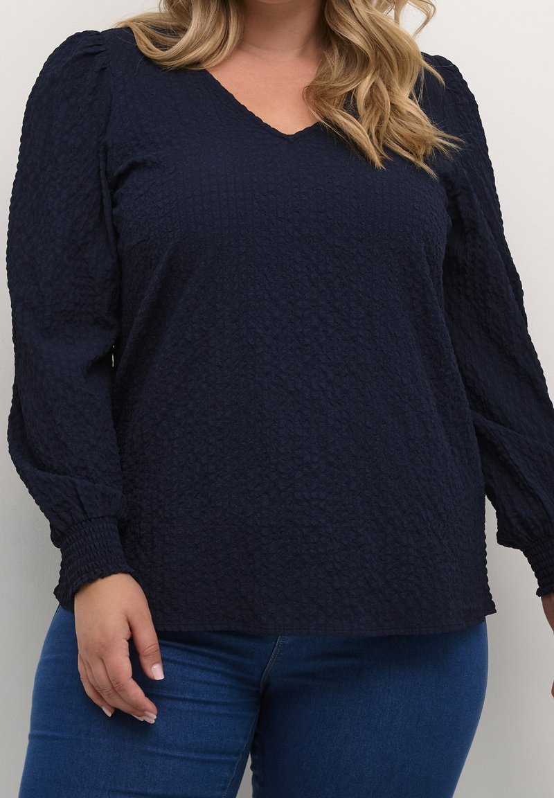 Femme portant un chemisier bleu marine texturé à manches longues avec poignets smockés et un jean bleu, debout devant un fond clair uni.