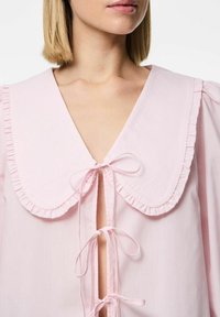 Camicia rosa con un ampio colletto arricciato, nastri anteriori e scollatura a V profonda. Realizzata in tessuto leggero, presenta maniche a palloncino e una superficie liscia.