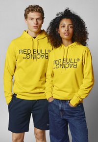 Geltoni džemperiai su juodu "RED BULL RACING" logotipu. Vyrų modelis dėvi šortus; moterų modelis vilki džinsus. Paprastas dizainas, be raštų.