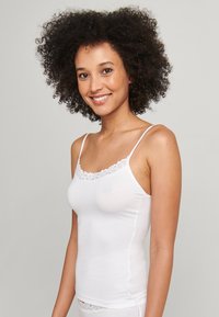 Vit camisole med spetskant, tunna band, slät textur, figurnära design, mjukt tyg, bärs av en person med lockigt hår, neutral bakgrund.