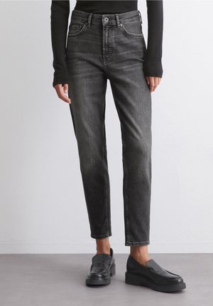 Person trägt ausgewaschene schwarze High-Waist-Jeans und schwarze Slip-On-Plateauschuhe und steht vor einem schlichten hellen Hintergrund.