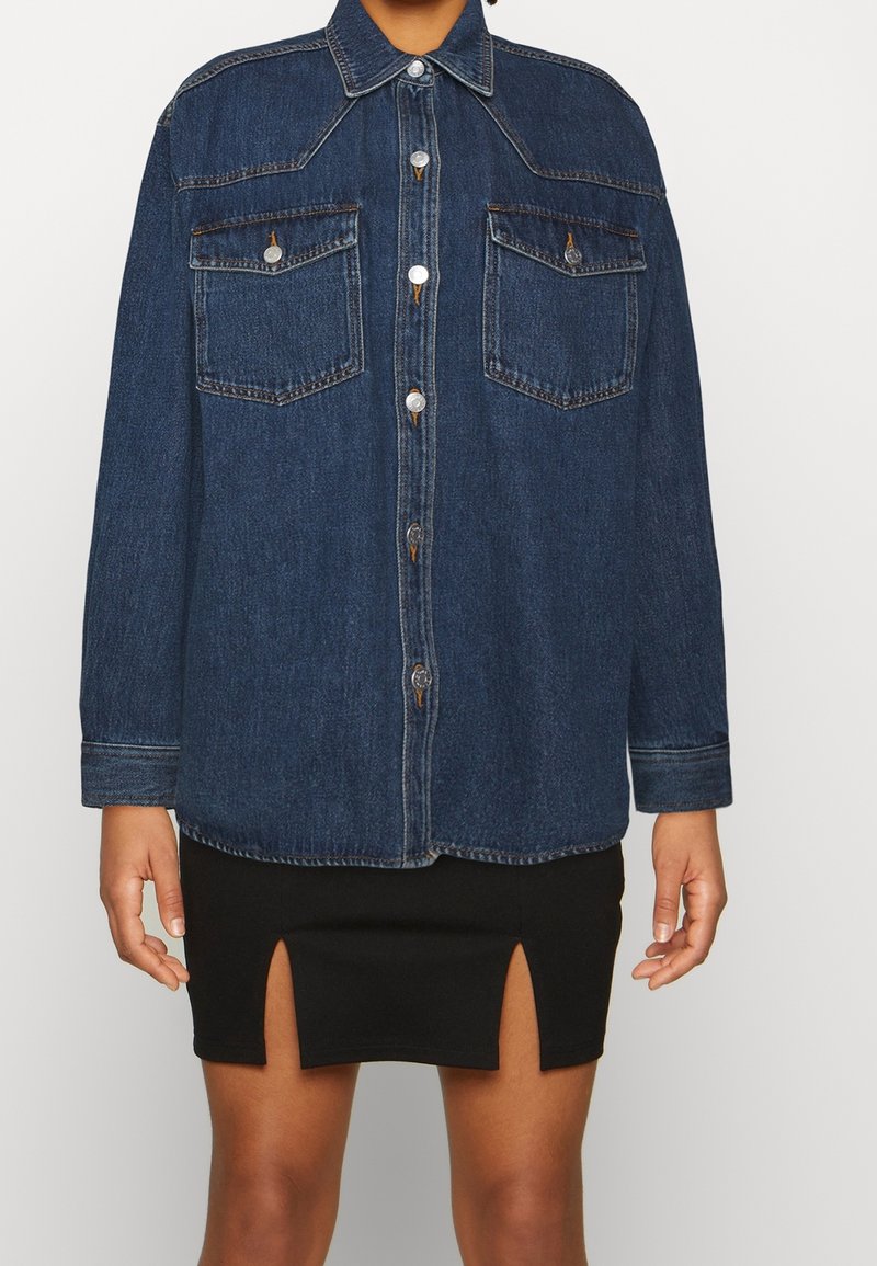 Denimskjorta i mörkblått med spetsig krage, knappföring fram, två bröstfickor och långa ärmar. Bärs över en svart kjol med slitsar.