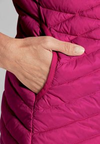 Gepostete Weste in lebhaftem Magenta, mit einer Seitentasche, in der eine Hand ruht, die die glatte Textur und die sichtbare Naht betont.