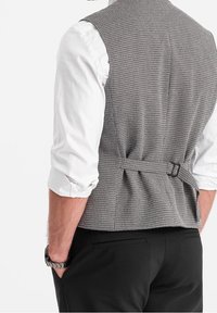 Grå texturerad väst med en figurnära design, baksidestrapsdetalj, buren över en vit långärmad skjorta, ihop med svarta byxor.