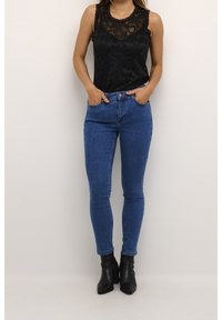 Svart spetslinne med rundad halsringning, i kombination med blå smala jeans. Modellen har på sig svarta ankelboots och står med händerna i fickorna.