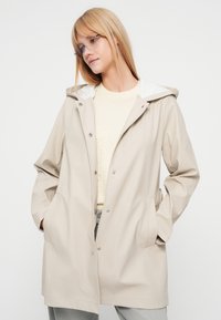 Veste imperméable beige avec capuche blanche, fermeture à boutons-pression, deux poches latérales, et une coupe décontractée, portée par-dessus un pull de couleur claire.