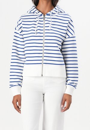 Vrouw die een wit zip-up hoodie met blauwe horizontale strepen en een witte broek draagt, staand tegen een effen achtergrond.