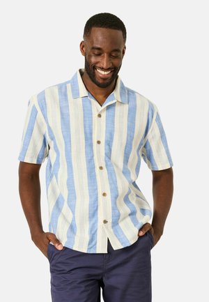 Man smiling with hands in pockets, wearing a short-sleeve blue and cream vertical striped button-up shirt and navy pants.