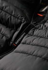 Czarna ocieplana kurtka z zamkiem. Posiada wyraźną metkę "SUPERDRY" w białym i czerwonym kolorze oraz gładką, błyszczącą fakturę.