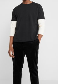 Schwarzes Kurzarm-T-Shirt mit langen cremefarbenen Ärmeln, strukturierter Stoff, Rundhalsausschnitt, getragen mit schwarzen Cordhosen. Keine sichtbaren Grafiken oder Muster.