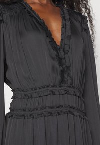 Robe noire à manches longues avec un profond décolleté en V et des détails à volants le long de l'encolure, de la taille et de la jupe, portée par une personne à la peau foncée.