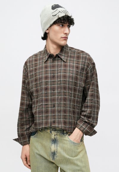 Brązowa koszula w kratę wykonana z lekkiego materiału, zapinana na guziki z przodu i długimi rękawami; w połączeniu z wyblakłymi zielonymi jeansami.