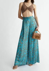 Un crop top en tricot marron associé à un pantalon large turquoise arborant un motif paisley marron. Le mannequin tient un sac à main beige avec des accents dorés.