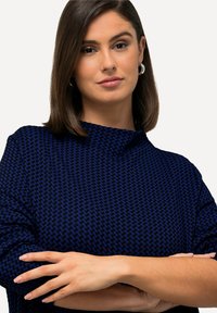 Top a fantasia blu navy con scollatura alta e tessuto testurizzato. Il modello incrocia le braccia, mettendo in mostra il design e la vestibilità del capo.