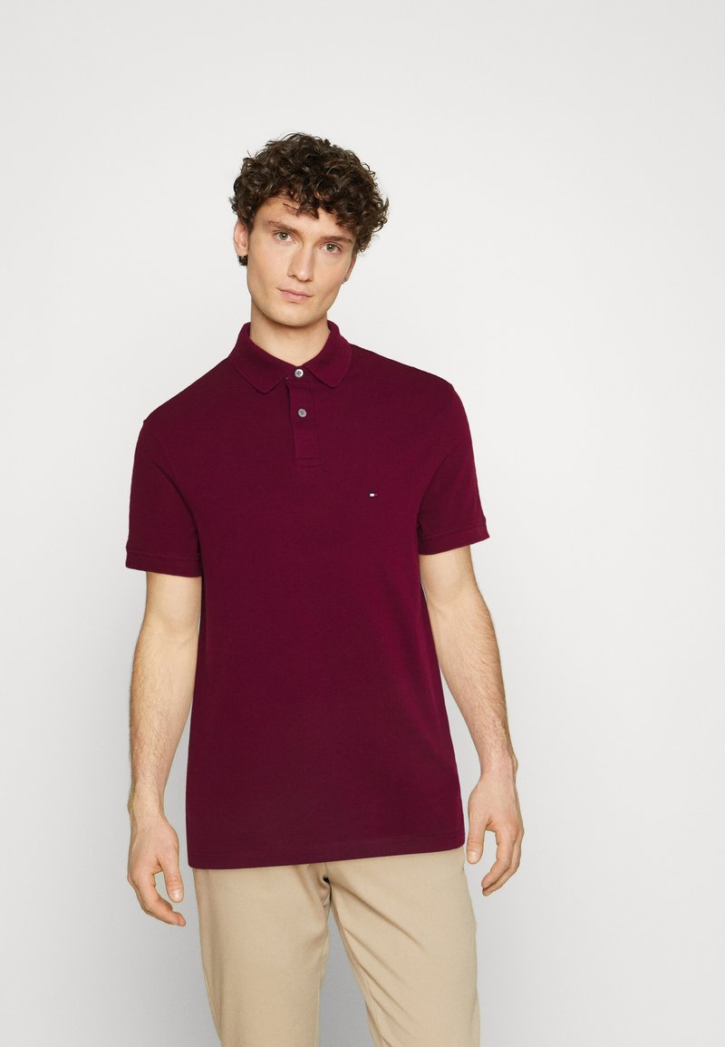 Polo bordeaux à manches courtes, avec patte de boutonnage deux boutons et un petit logo sur le côté gauche de la poitrine, associé à un pantalon beige clair.