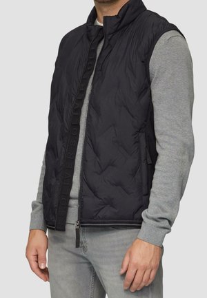 Mand iført en sort quiltet vest uden ærmer over en grå langærmet sweater og lysegrå jeans, stående foran en ensfarvet baggrund.