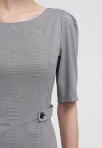 Robe en pied-de-poule noir et blanc avec des manches courtes bouffantes, un col rond et des détails de boutons à la taille. Texture lisse.