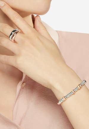 Hand mit zweifarbigen Silber- und Roségoldring nahe am Hals, trägt ein Armband mit abwechselnden Silber- und Roségoldperlen, an einer Person in einem rosa Hemd.
