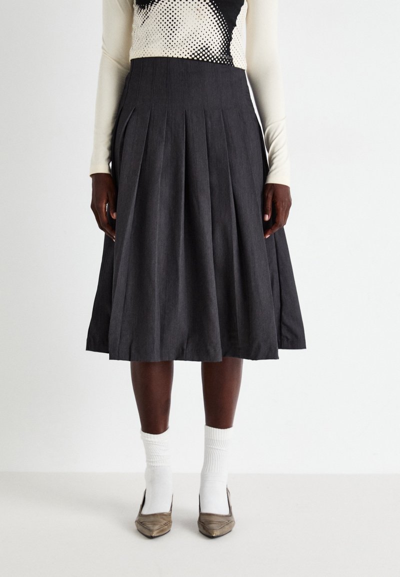 Personne portant une jupe midi plissée noire, un haut blanc à manches longues, des chaussettes blanches et des chaussures pointues bronze, debout devant un fond uni.
