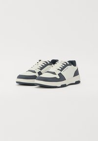 Zapatillas bajas blancas y marinas con cordones en la parte delantera y logo de la marca en un lado, posicionadas sobre un fondo claro y liso.
