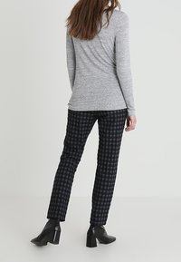 En kvinna står med ryggen mot kameran iförd en grå långärmad tröja, svart och grå rutiga byxor samt svarta klackade ankelstövlar framför en neutral bakgrund.