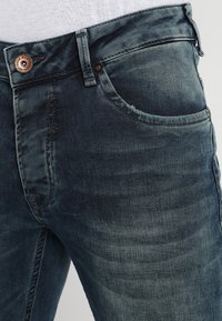 Jeans de mezclilla azul oscuro con una textura ligeramente desgastada, que presentan un botón de cobre, aros para el cinturón y un diseño estándar de cinco bolsillos.