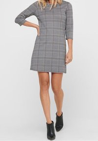 Graues Karo-Kleid mit Dreiviertelärmeln, A-Linien-Schnitt und Seitentaschen. Kombiniert mit schwarzen Knöchelstiefeln. Glatte Textur, schlichtes Design.