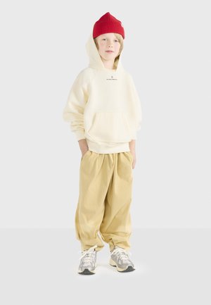 Crèmekleurige hoodie met een voorzak, beige loszittende broek en grijze sneakers. Het kind draagt een rode gebreide muts.