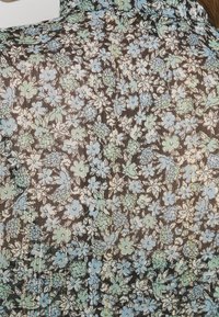 Tejido con estampado floral que presenta pequeñas flores azules y verdes sobre un fondo oscuro, con sutiles acentos metálicos y una textura transparente.