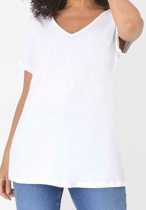 Kvinde iført en løs hvid kortærmet V-hals top med oprullede ærmekanter, kombineret med blå jeans mod en ensfarvet baggrund.